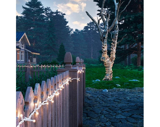 Warm white LED string lights wrapped around a tree and draped along a wooden fence at dusk.