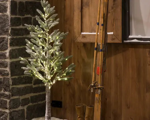 96-light LED snowy pine tree standing next to vintage skis and boots against a wood-paneled wall.