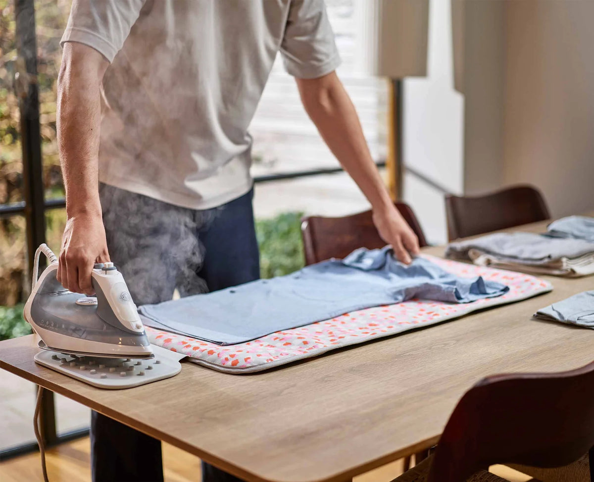 Person ironing a blue shirt on a table using the Joseph Swift Ironing/Steaming Mat in peach blossom pattern