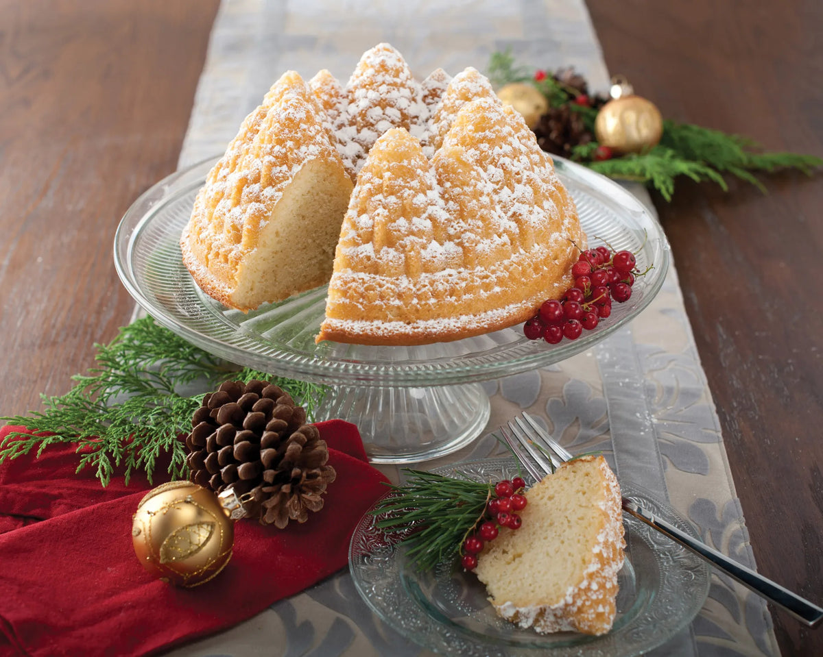 Bundt cake shaped like pine trees dusted with powdered sugar on a glass cake stand with holiday decor.