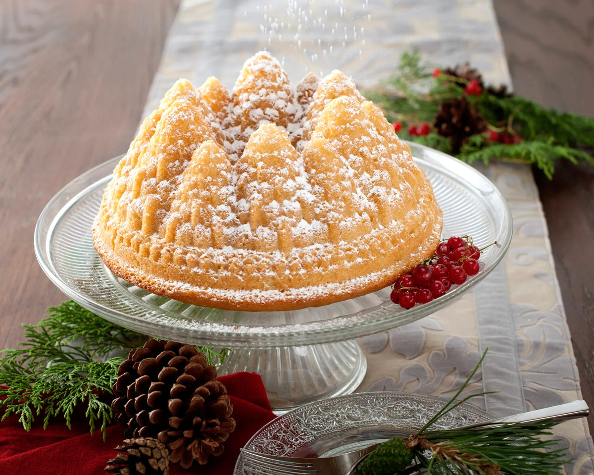 Bundt cake shaped like pine trees dusted with powdered sugar on a glass cake stand with holiday decor.