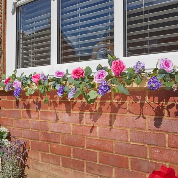Eden Blooms Garland 180cm with pink and purple flowers draped along a brick wall under a window.