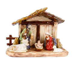 A ceramic nativity scene figurine set featuring Mary, Joseph, baby Jesus in a manger, a cow, and a sheep, arranged inside a rustic stable with a brown roof on a white background.