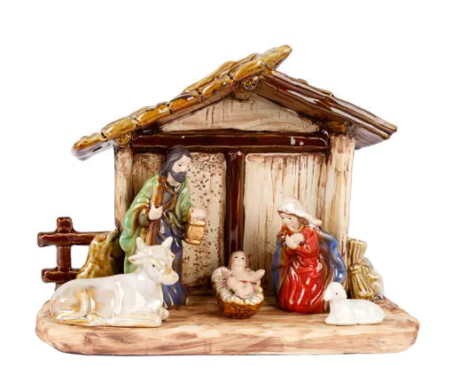 A ceramic nativity scene figurine set featuring Mary, Joseph, baby Jesus in a manger, a cow, and a sheep, arranged inside a rustic stable with a brown roof on a white background.