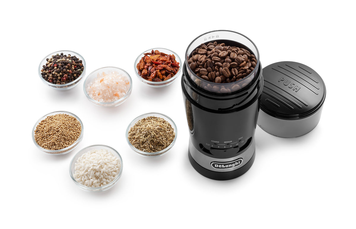 Delonghi Electric Blade Coffee Grinder filled with coffee beans, surrounded by bowls of spices and grains on white