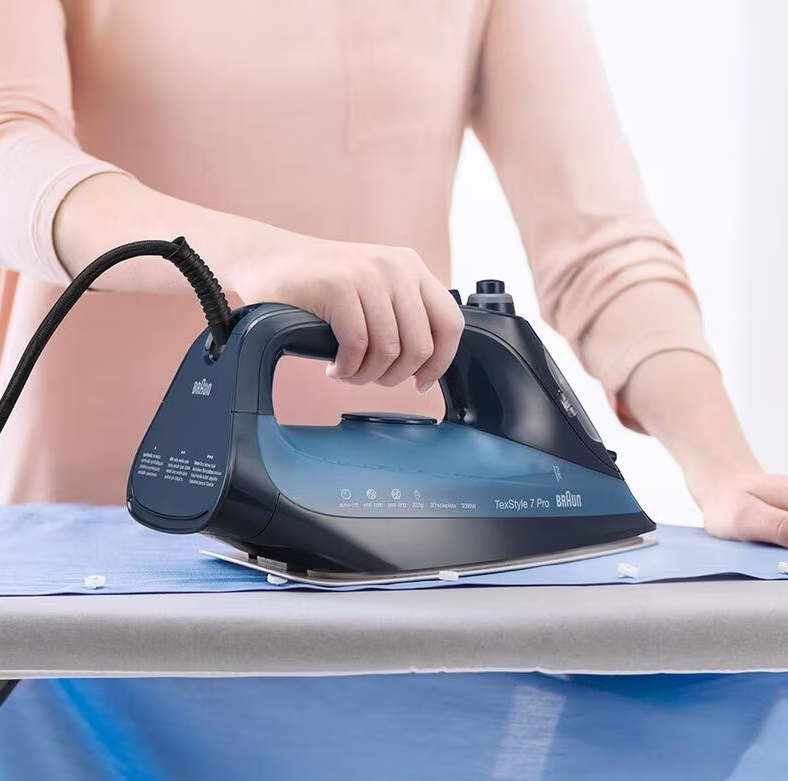 Person using a blue and black Braun Steam Generator Iron SI7160BL on a light blue shirt on an ironing board.