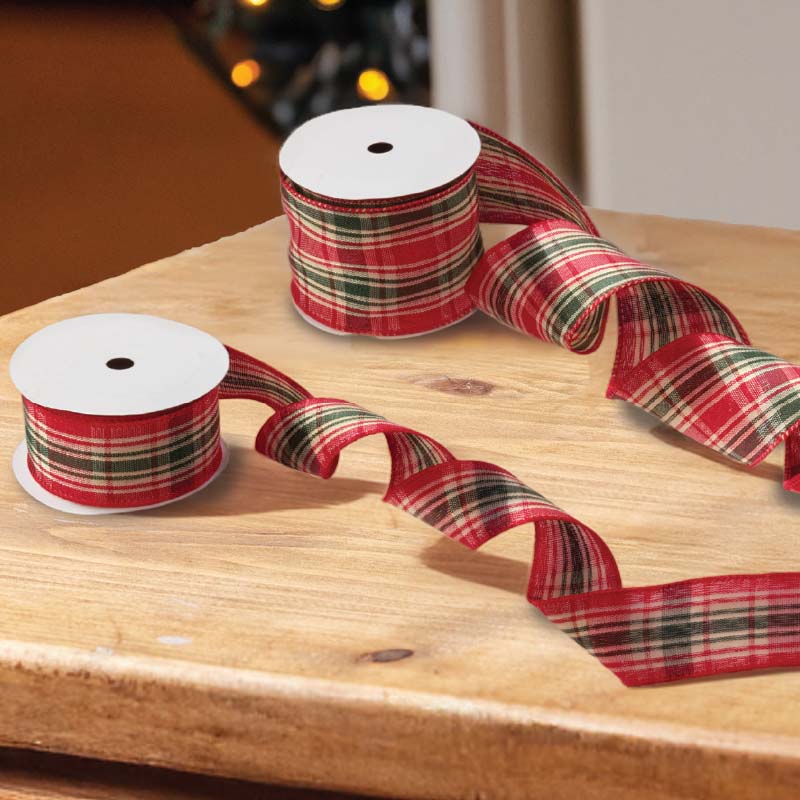 Three spools of holiday ribbon with a red, green, and black plaid pattern, displayed on a wooden tabletop with festive blurred lights in the background.