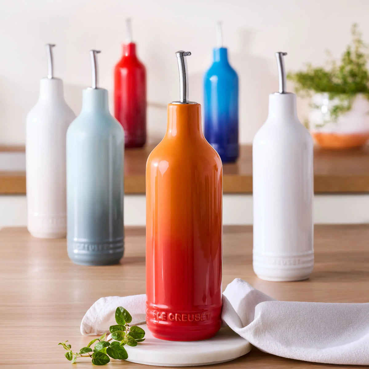 Le Creuset oil cruet in red-orange gradient with metal pour spout on wooden table with herbs and cloth.