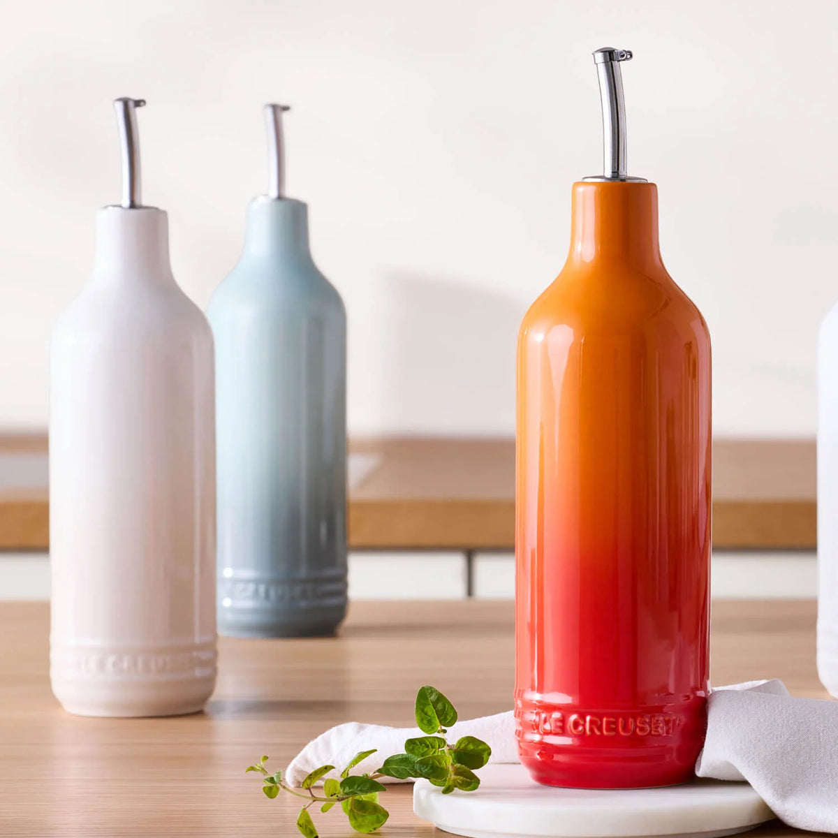 Le Creuset Oil Cruet in volcanic red-orange with stainless steel pourer on kitchen counter with herbs and cloth.