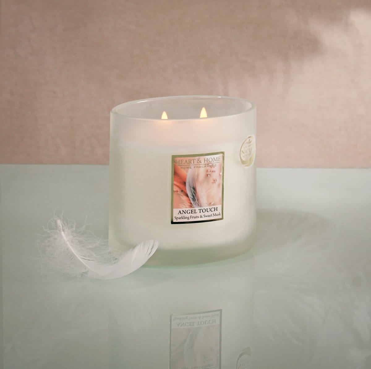 Twin wick Angel Touch candle in frosted glass with lit flames and a white feather beside it on a reflective surface.
