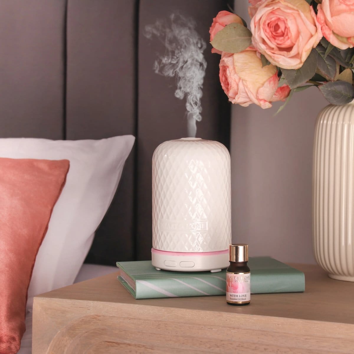 White ceramic diffuser emitting mist beside a 10ml With Love essential oil bottle on a bedside table.