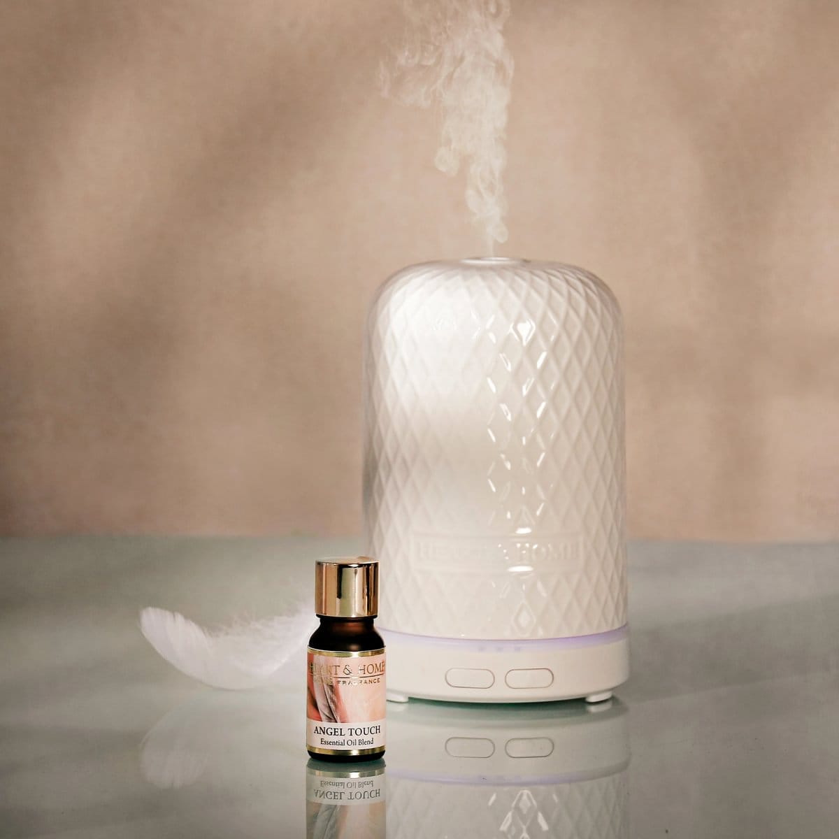 Angel Touch essential oil bottle next to a white ceramic diffuser releasing mist on a reflective surface.