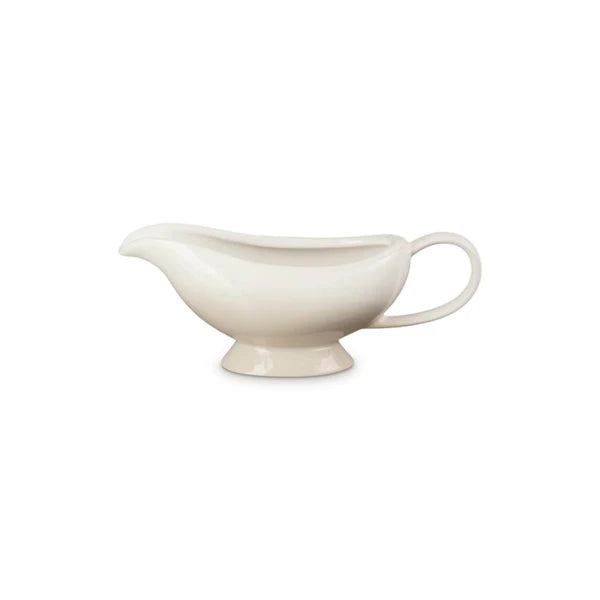 Le Creuset Heritage gravy boat in meringue color with a curved spout and handle on a pedestal base.