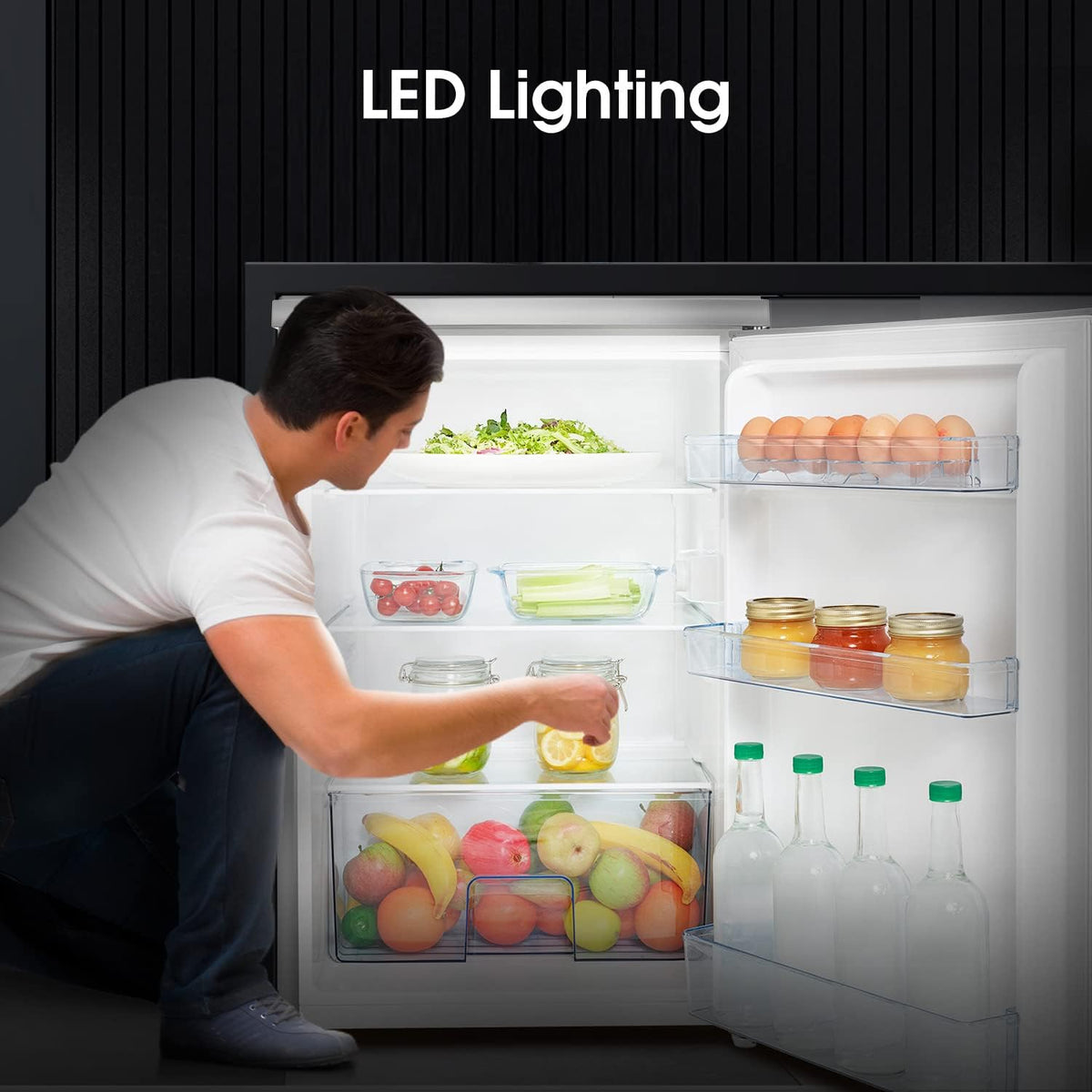 Man placing a jar inside a Hisense 56cm undercounter fridge with LED lighting and stocked shelves.