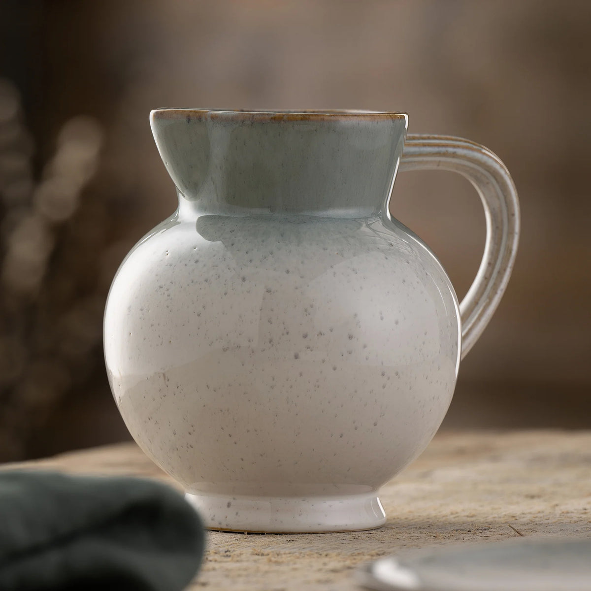Close-up of a speckled ceramic Tivoli Pitcher with a curved handle and wide spout on a wooden surface.