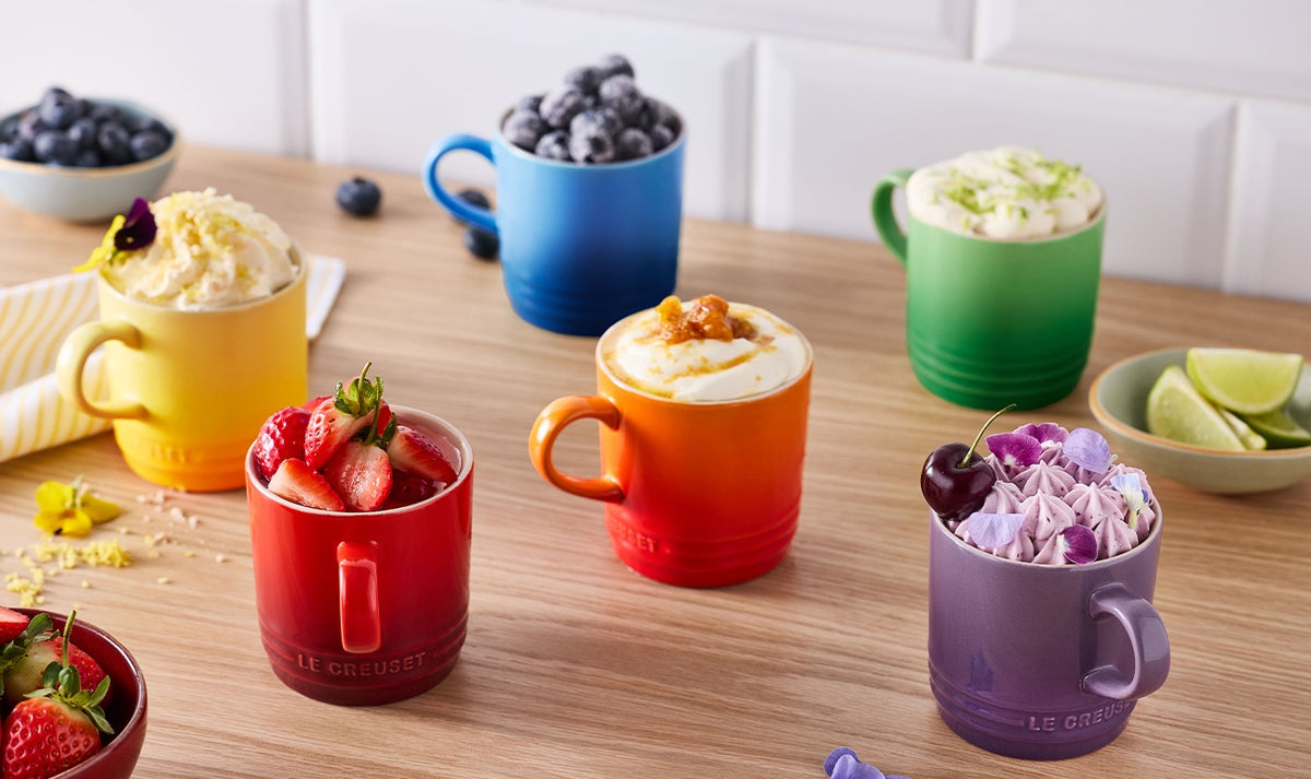 Six colorful Le Creuset cappuccino mugs filled with fruit, whipped cream, and toppings on a wooden table.
