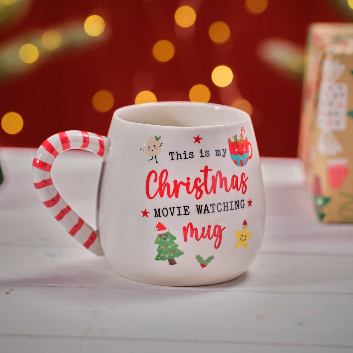 White ceramic Christmas movie mug with candy cane handle and festive illustrations on a wooden surface.