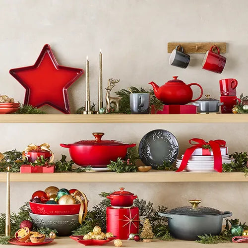 Le Creuset Stoneware Holly Plate 19cm Thyme in green on a wooden shelf with festive decor and cookware.