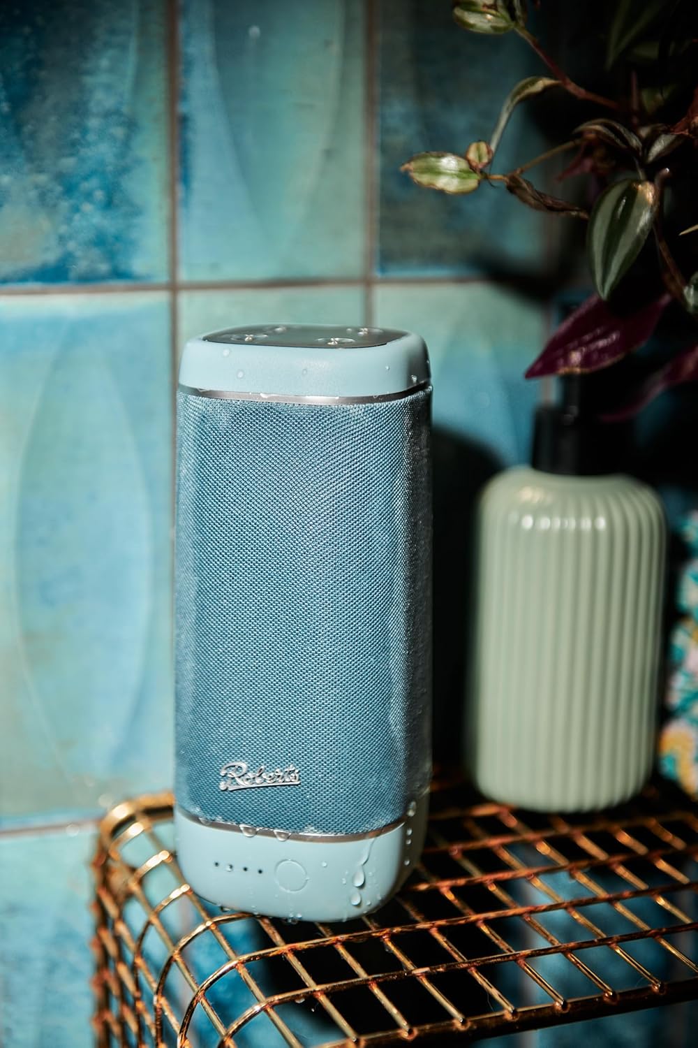 Roberts REUNIONDE portable Bluetooth speaker in duck egg blue with water droplets on a metal rack.