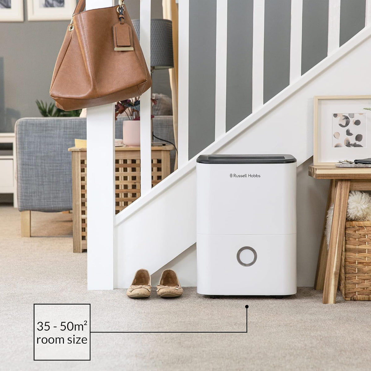 Russell Hobbs 20L dehumidifier placed under stairs in a living room with shoes nearby and a brown bag hanging on railing.