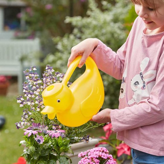 Child holding a yellow DuckCan kids watering can, watering flowers in a garden setting.