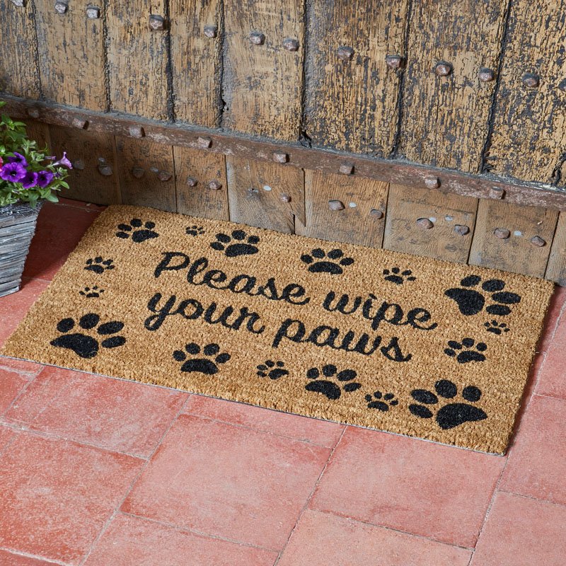 Rectangular brown doormat with black paw prints and "Please wipe your paws" text on red tiled floor by wooden door.