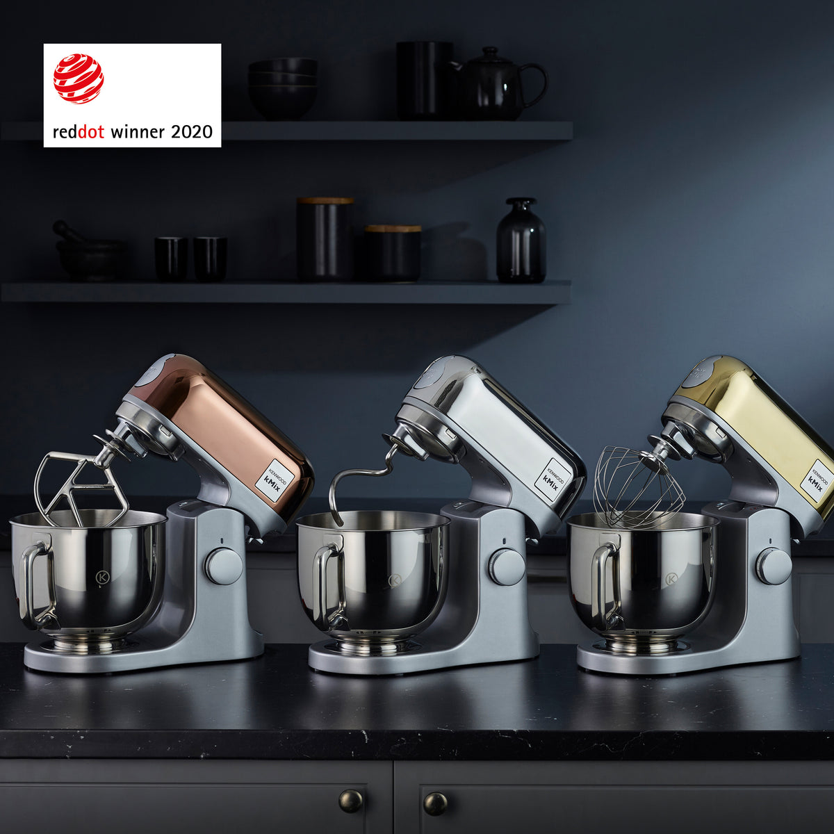 Three Kenwood kMix kitchen mixers in rose gold, silver, and gold finishes on a dark countertop.