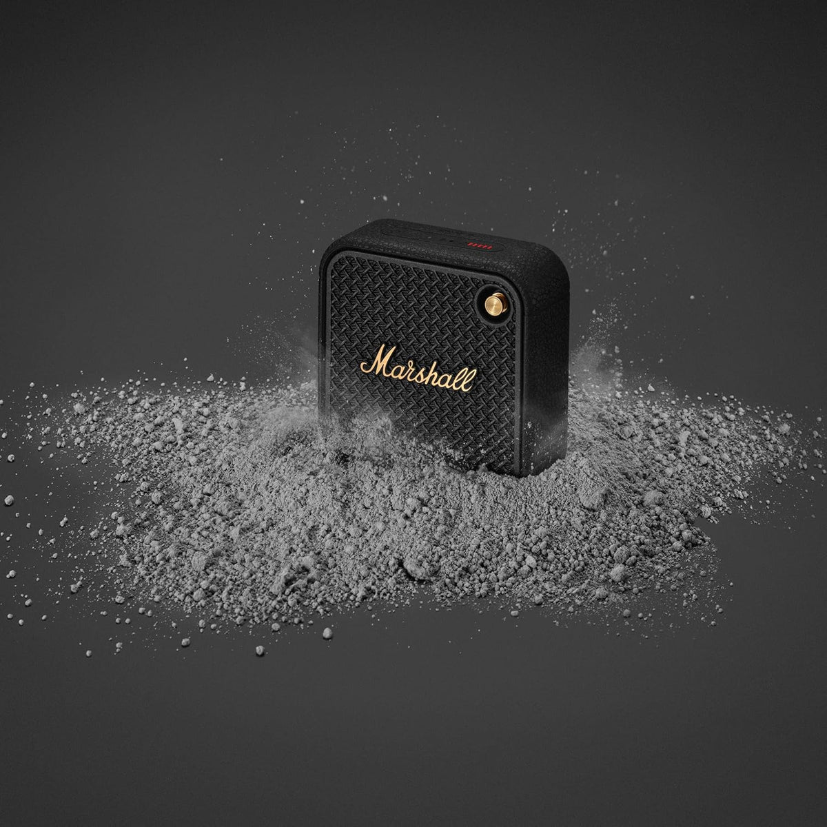 Marshall Willen II Black & Brass portable speaker partially buried in gray dust on a dark background