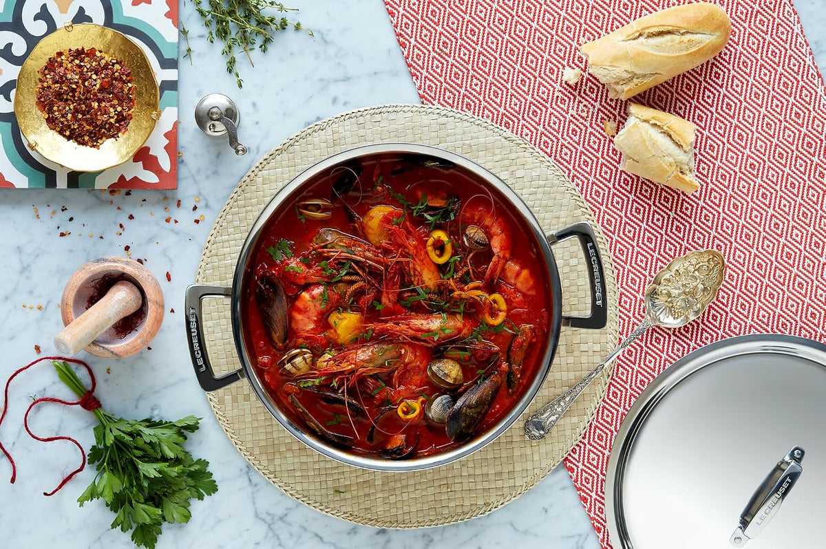 Le Creuset stainless steel deep casserole filled with seafood stew, shown with lid and bread on marble surface.