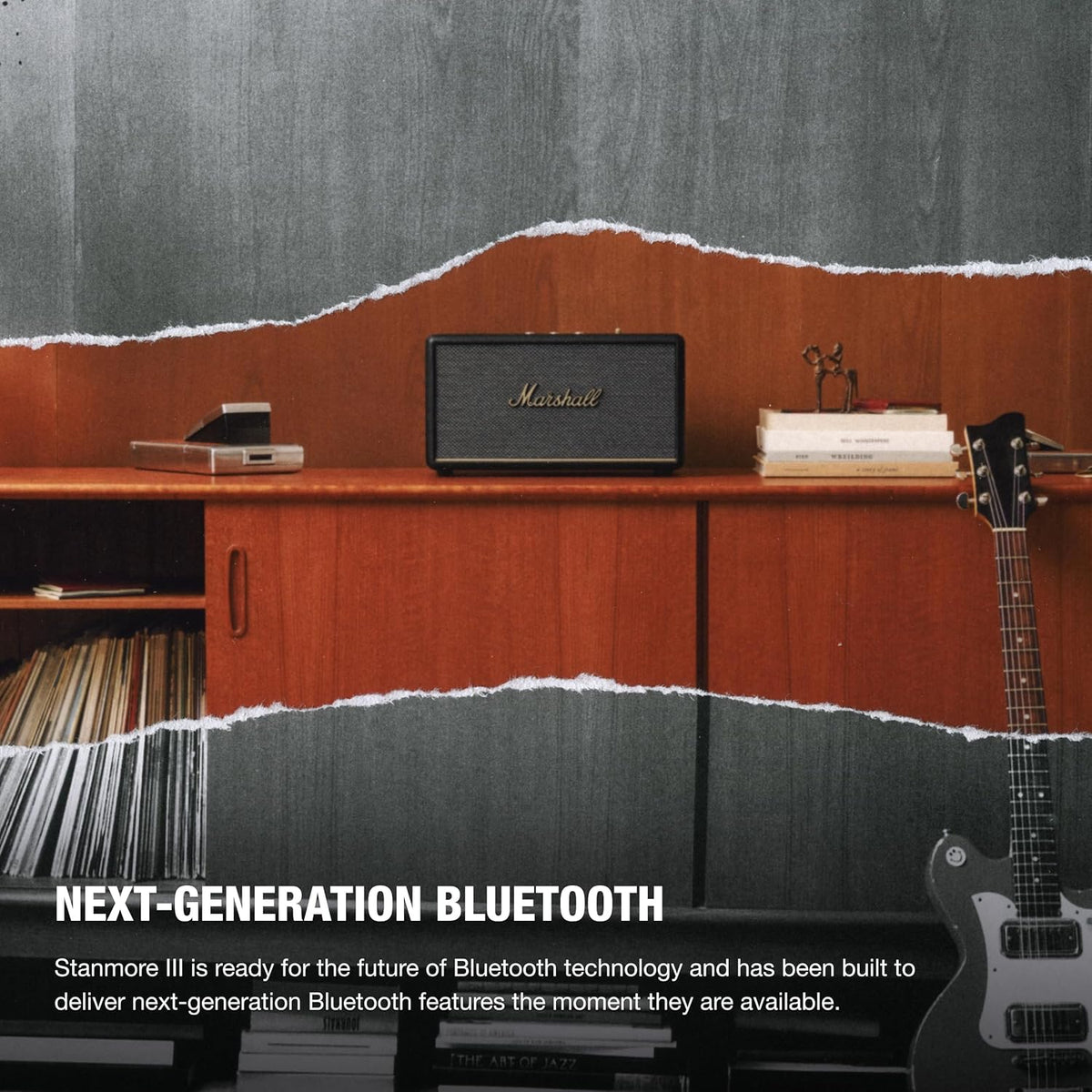 Marshall Stanmore III Bluetooth speaker in black on a wooden cabinet with books and a guitar nearby