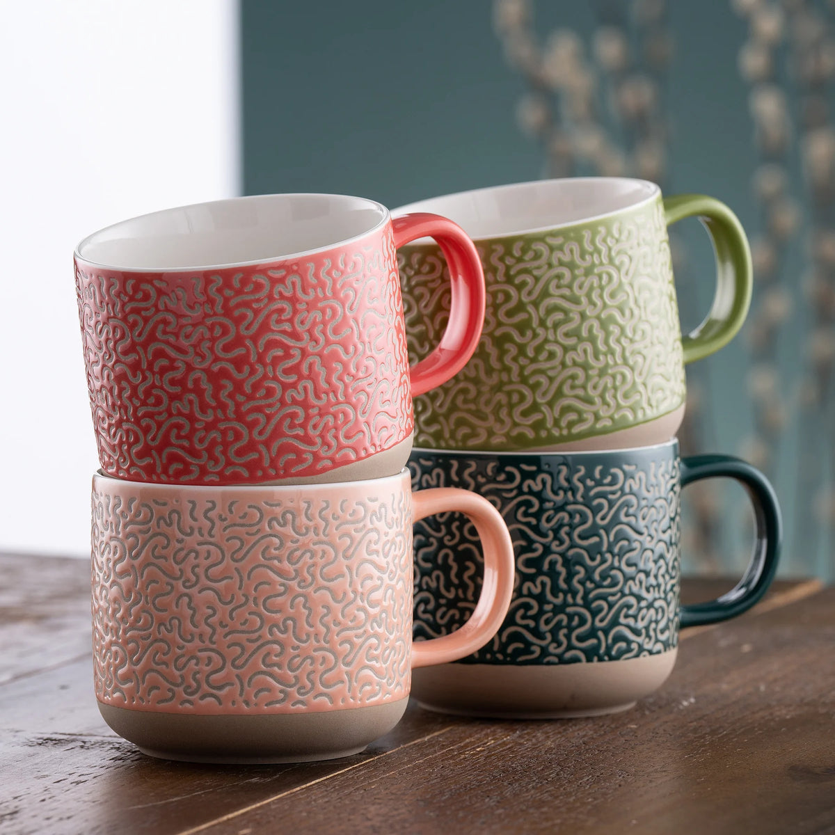 Four ceramic Squiggle Mugs in coral, pink, green, and dark teal stacked on a wooden surface.