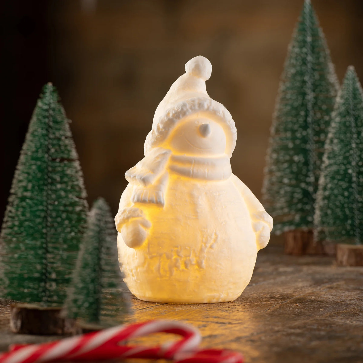 Belleek Snowman LED glowing softly on wooden surface with miniature Christmas trees in background