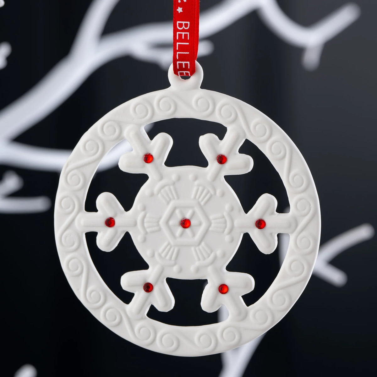 White Belleek snowflake ornament with red gem accents and a red hanging ribbon against a dark background.