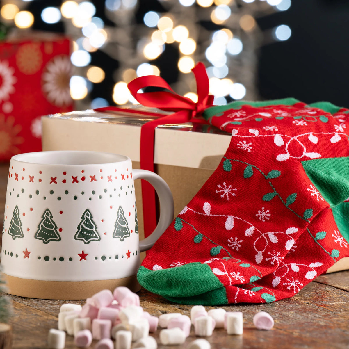 Festive Belleek mug with Christmas tree pattern, red and green holiday socks, gift box, and marshmallows on wood table.