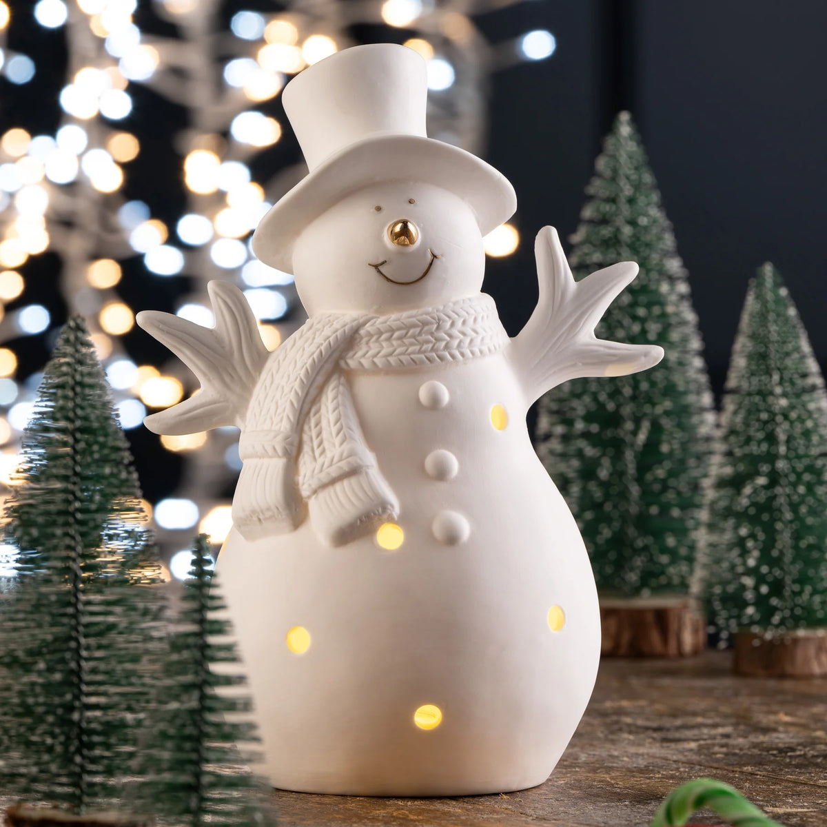 White ceramic Belleek Snowman LED with gold nose and scarf, surrounded by miniature Christmas trees and lights.