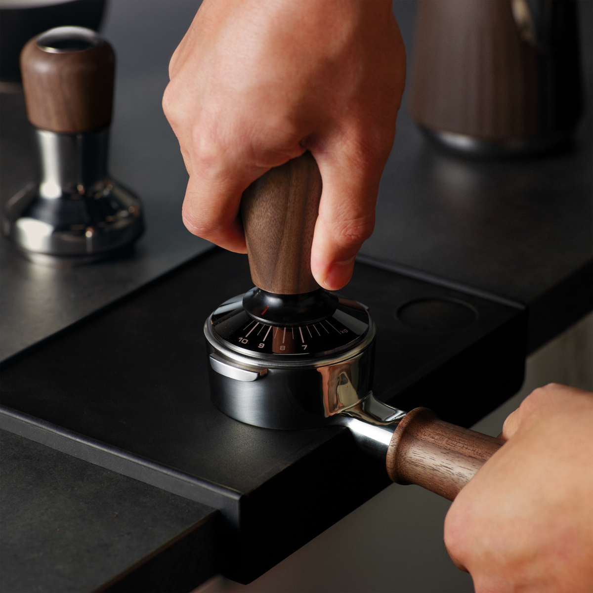 Hand pressing the Sage the Force Gauge Tamper with a wooden handle onto a portafilter on a black surface.