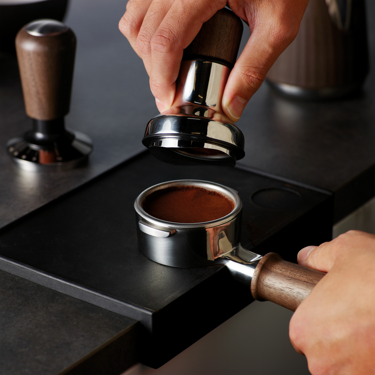 Hand pressing coffee grounds with Sage the Distribution Duo 58mm tamper on black tray and portafilter.