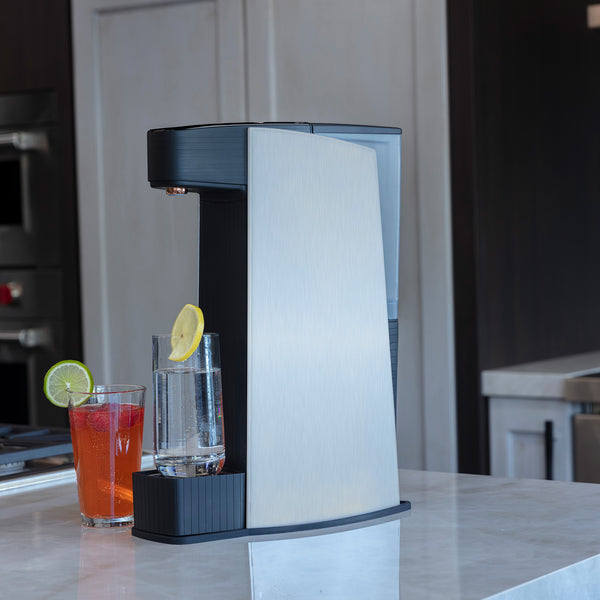 Carbon8 One Touch Sparkling Water Dispenser with two glasses, one with lemon and one with lime garnish on a kitchen counter