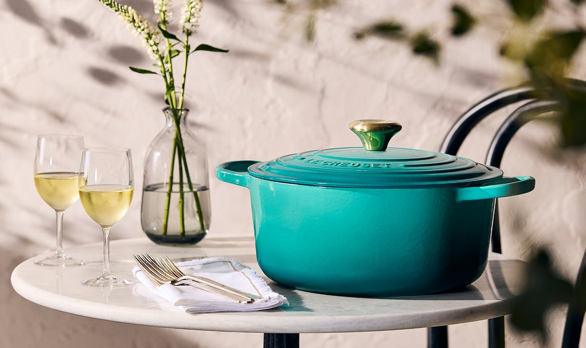Le Creuset 28cm blue Riviera round casserole with lid on a white table beside wine glasses and cutlery