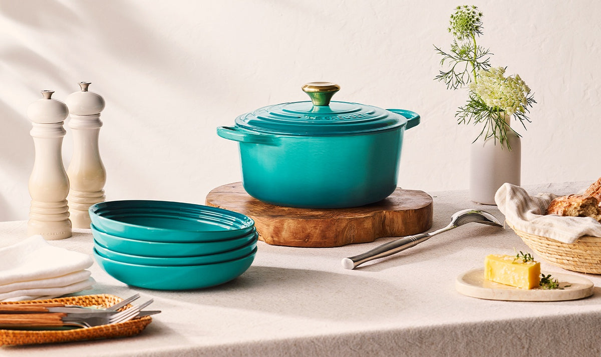 Le Creuset 28cm blue Riviera round casserole with lid on wooden board, surrounded by plates and tableware.