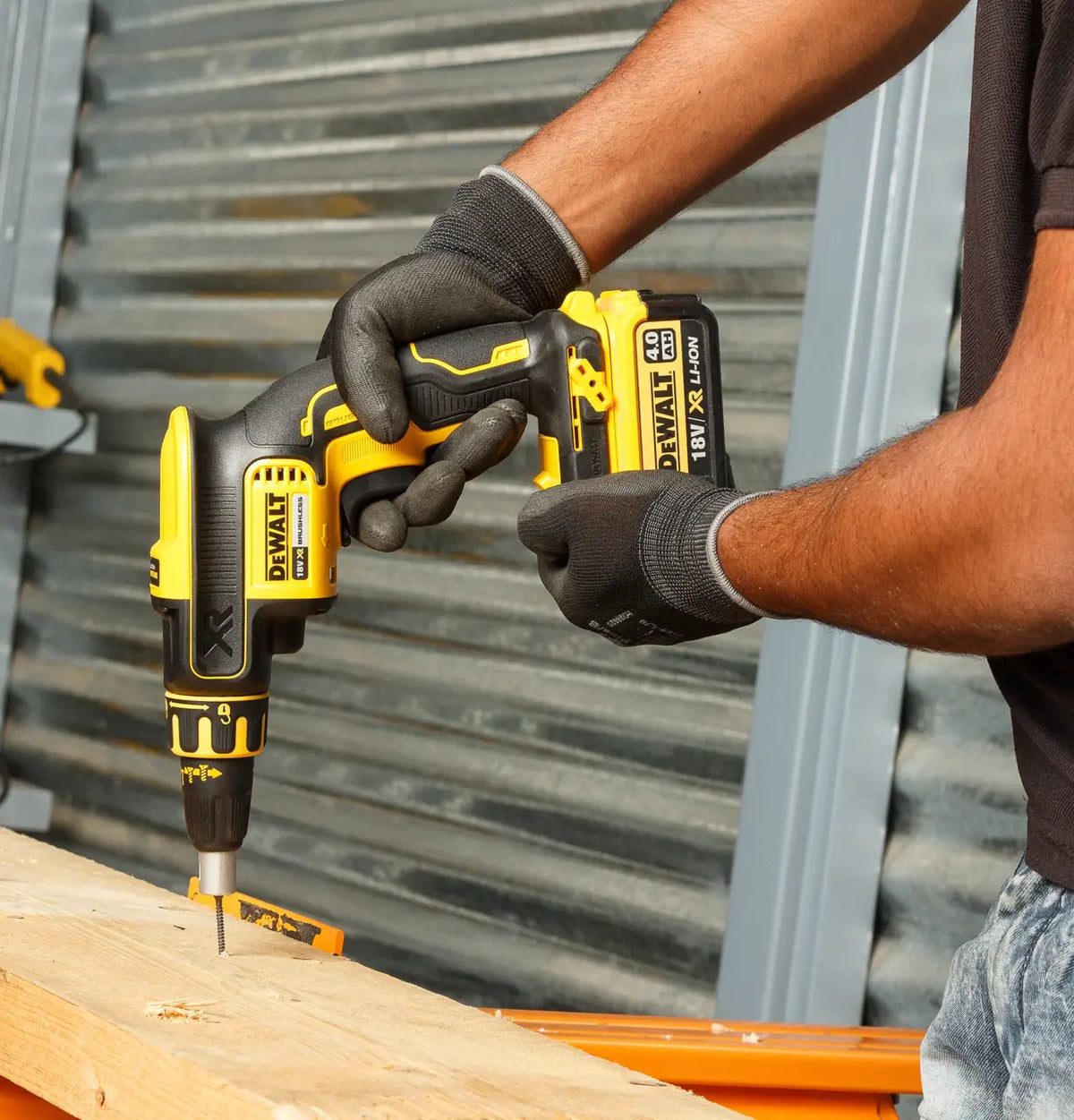 Hands wearing black gloves using a Dewalt 18V drywall screwdriver driving a screw into wood.