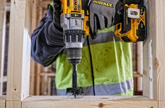 DeWalt self-feed drill bit attached to a drill, drilling into a wooden frame held by a worker in a green vest.