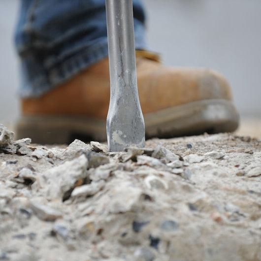 DeWalt SDS Plus steel flat chisel breaking concrete with a worker's boot in the background.