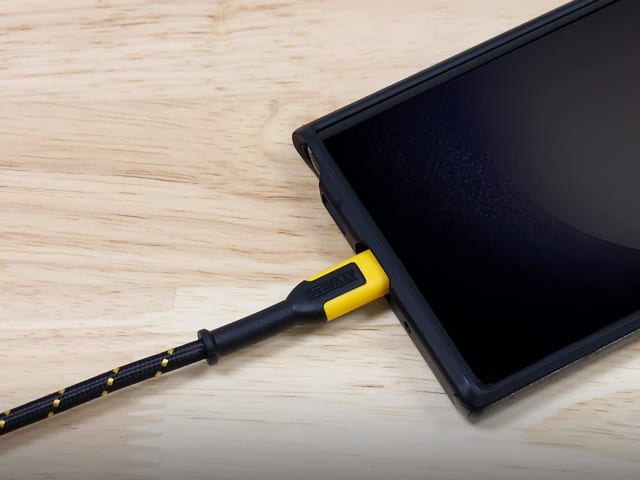 Close-up of DEWALT USB A to USB C reinforced charging cable plugged into a smartphone on a wooden surface.