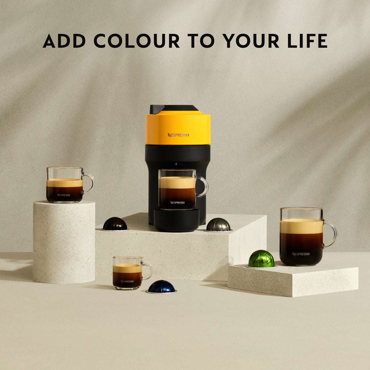 Delonghi Nespresso Vertuo Pop coffee machine in black with yellow top, surrounded by coffee capsules and glass cups of