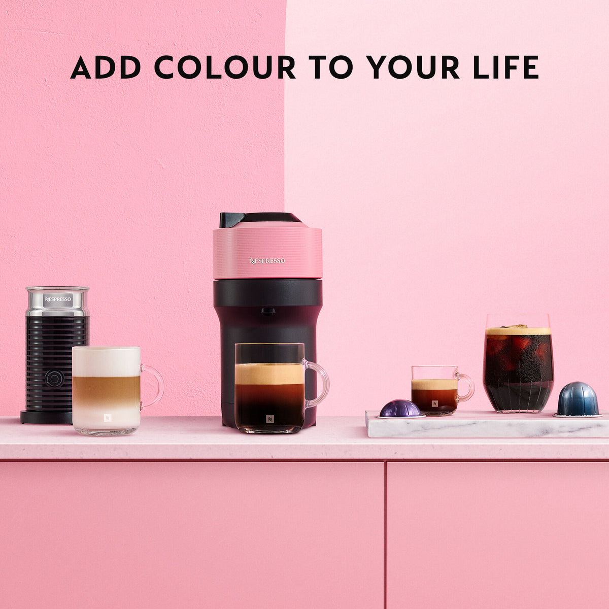 Delonghi Nespresso Vertuo Pop coffee machine in pink with milk frother, coffee cups, and capsules on pink counter