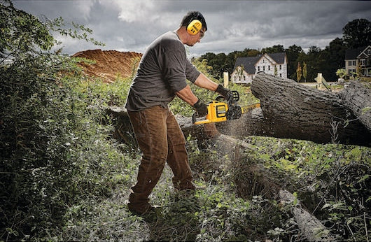 Man wearing yellow DeWalt DPG17 Bluetooth hearing protectors while cutting a large log with a chainsaw outdoors