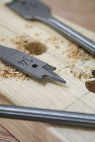 Close-up of flat drill bits from the 13-piece Flat Bit Set on a wooden board with drilled holes and wood shavings.