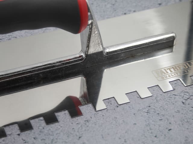 Close-up of the notched edge and handle of the FAI Full Notched Trowel 13" on a textured surface.