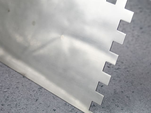 Close-up of the notched edge of the 13" FAI Full Notched Trowel on a gray surface.